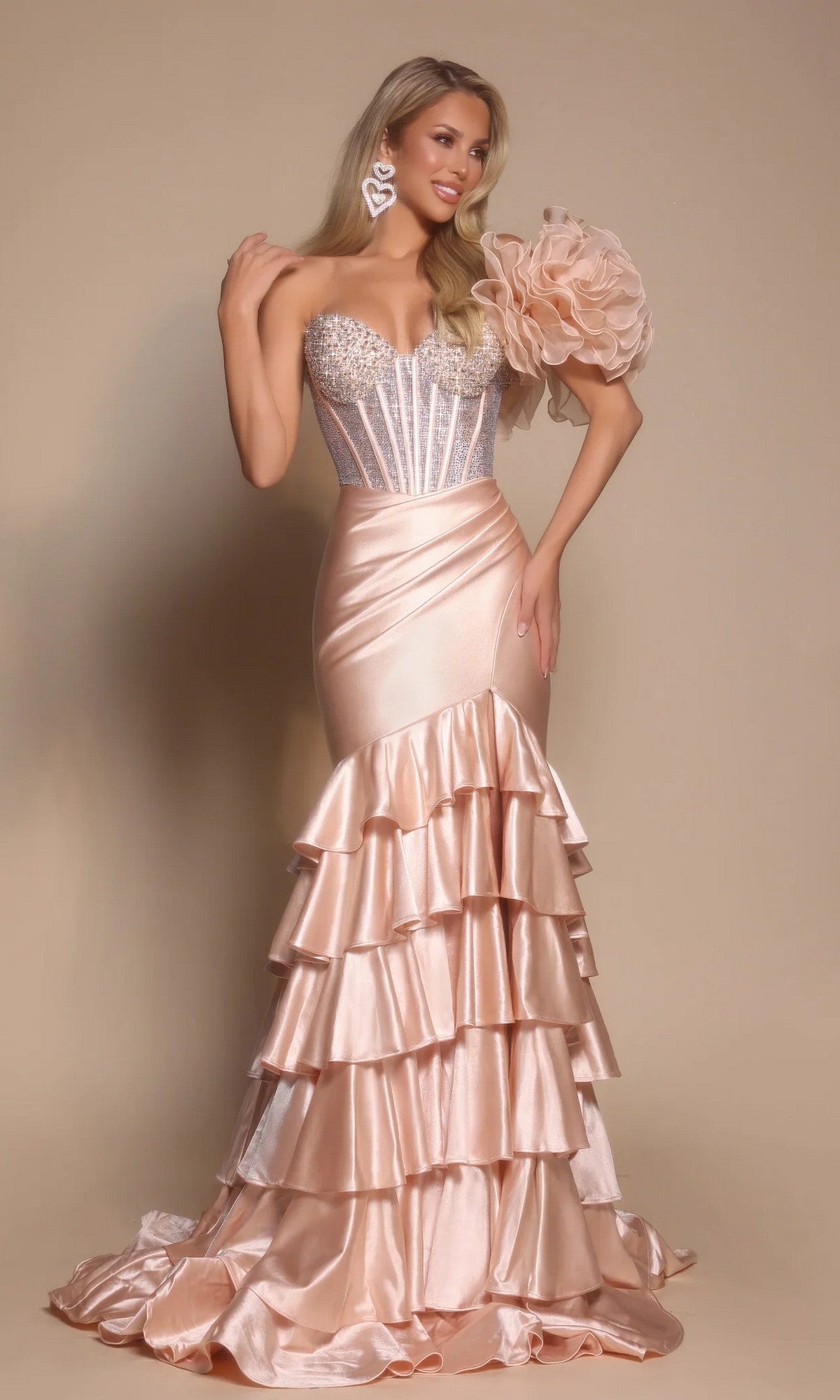 A woman poses in the Portia and Scarlett PS26141 long formal dress in champagne, featuring a strapless beaded corset bodice, large flower accent on one shoulder, and a tiered satin mermaid skirt against a beige background.